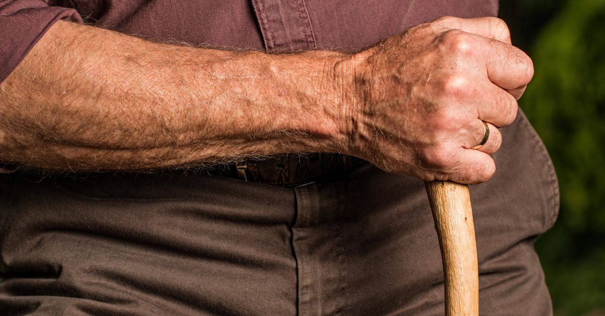 Close-up of a senior adult's hand gripping a wooden walking cane outdoors, symbolizing support and aging.