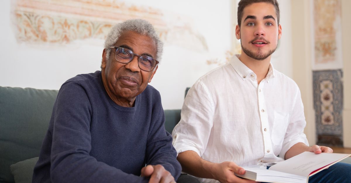 Elderly man and young volunteer sharing a moment reading indoors, promoting social assistance.