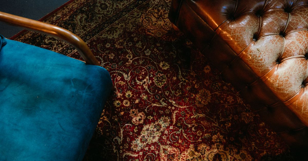 Elegant scene of a vintage rug, blue velvet chair, and leather sofa in warm lighting.