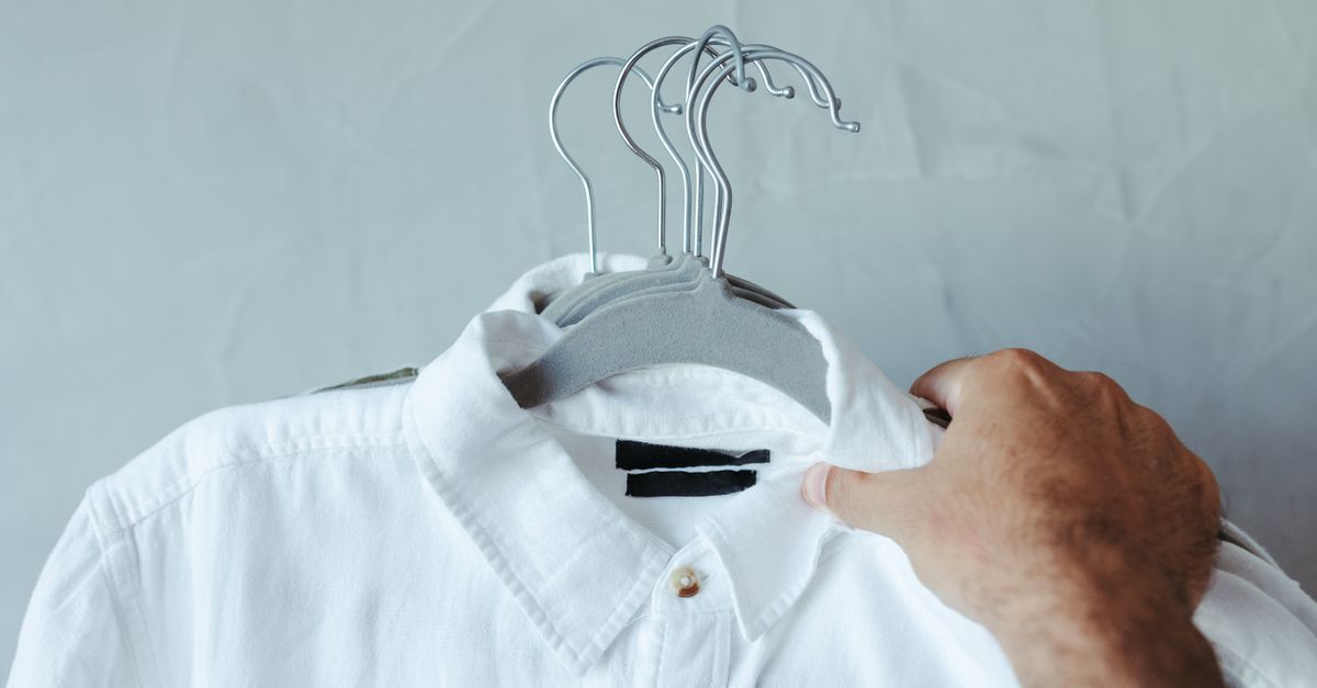 Close-up of a white shirt on a hanger held by a hand against a plain background.