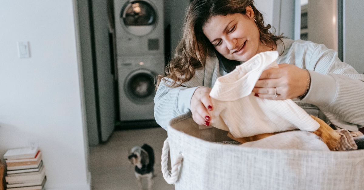 Optimistic female with clean clothes in basket having phone conversation while standing in light washroom with washing machines and dog