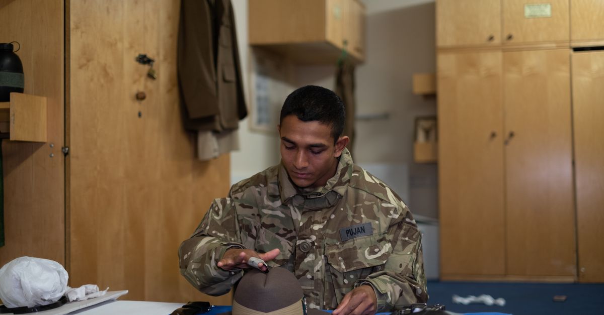 A soldier in camouflage uniform focuses on polishing a hat inside a barracks room.