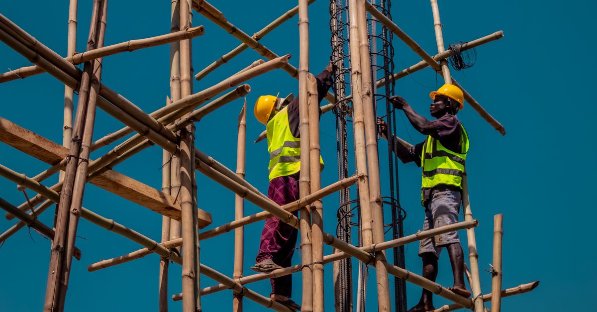 Two construction workers on bamboo scaffolding, wearing safety gear under a clear blue sky.