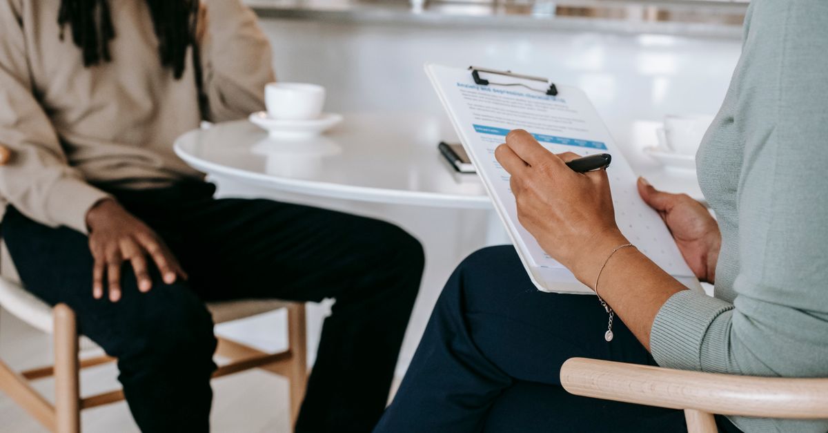 Crop unrecognizable female psychotherapist or interviewer asking questions and taking notes in clipboard during interview with black male