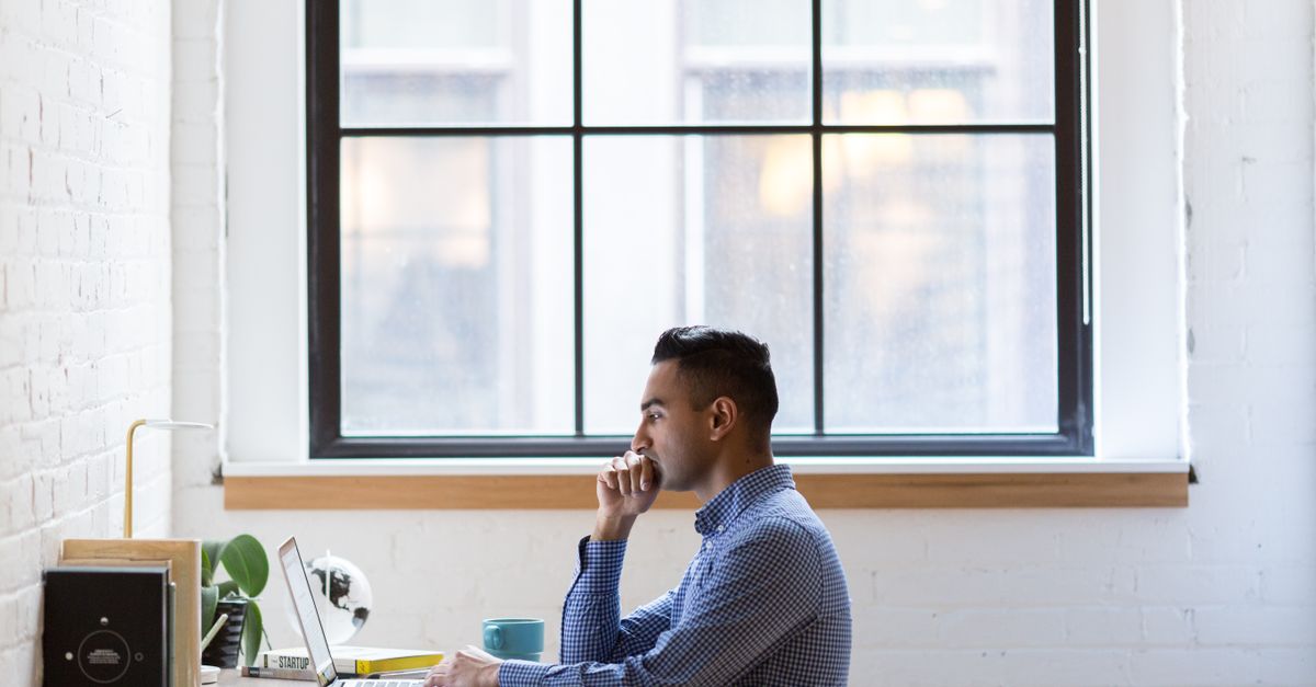 Focused male professional working on a laptop at an office desk by a large window.