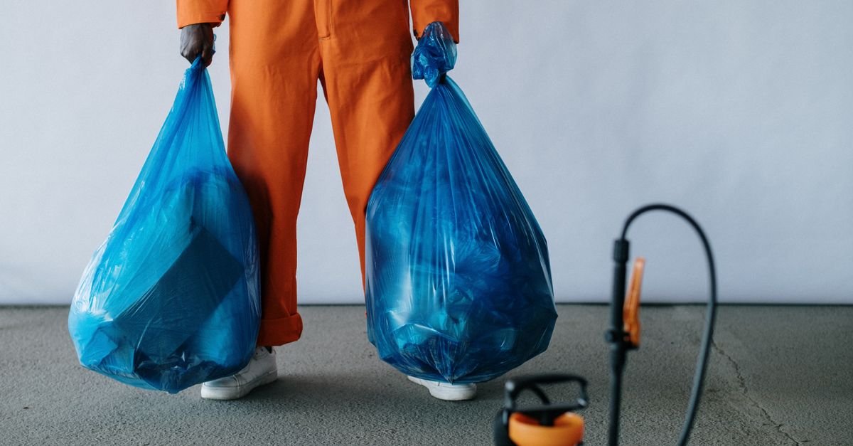 Sanitation worker in orange overalls holding blue trash bags indoors for waste management.