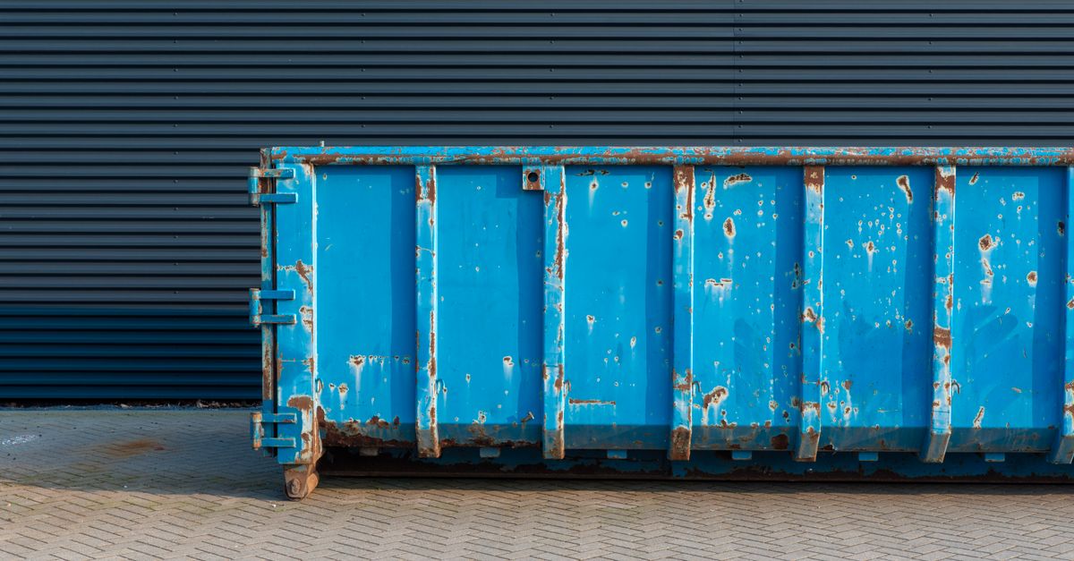 A blue rusty industrial container sits against a corrugated metal wall on a paved surface.