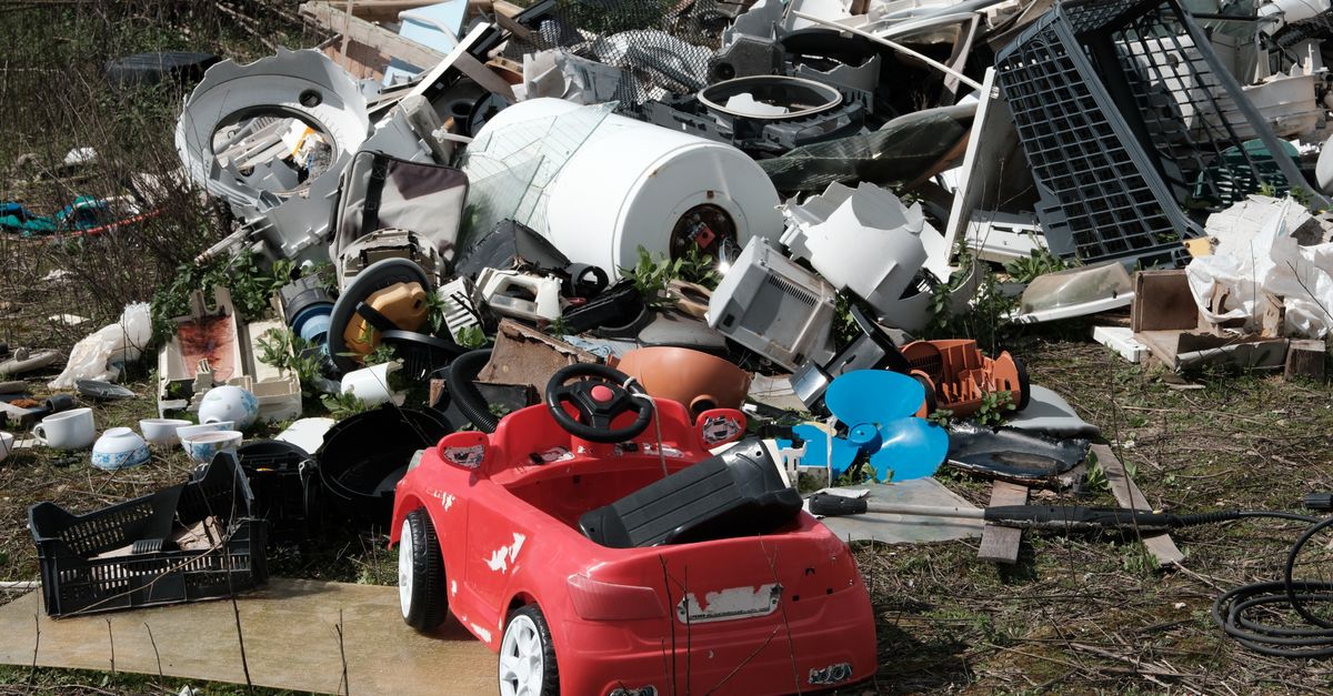 A red toy car sits among discarded appliances and debris, symbolizing environmental neglect.