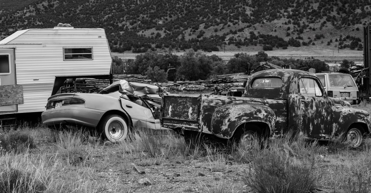 Black and white photo of rusty abandoned vehicles and camper in rural setting.