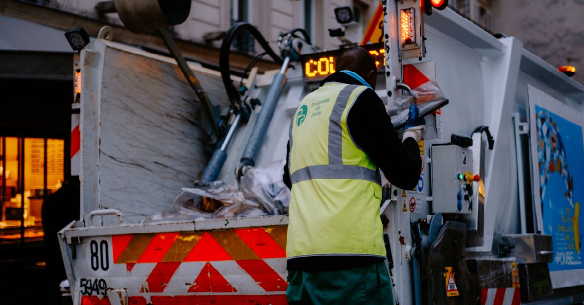 A garbage collector works on a city street, managing waste collection at dusk.