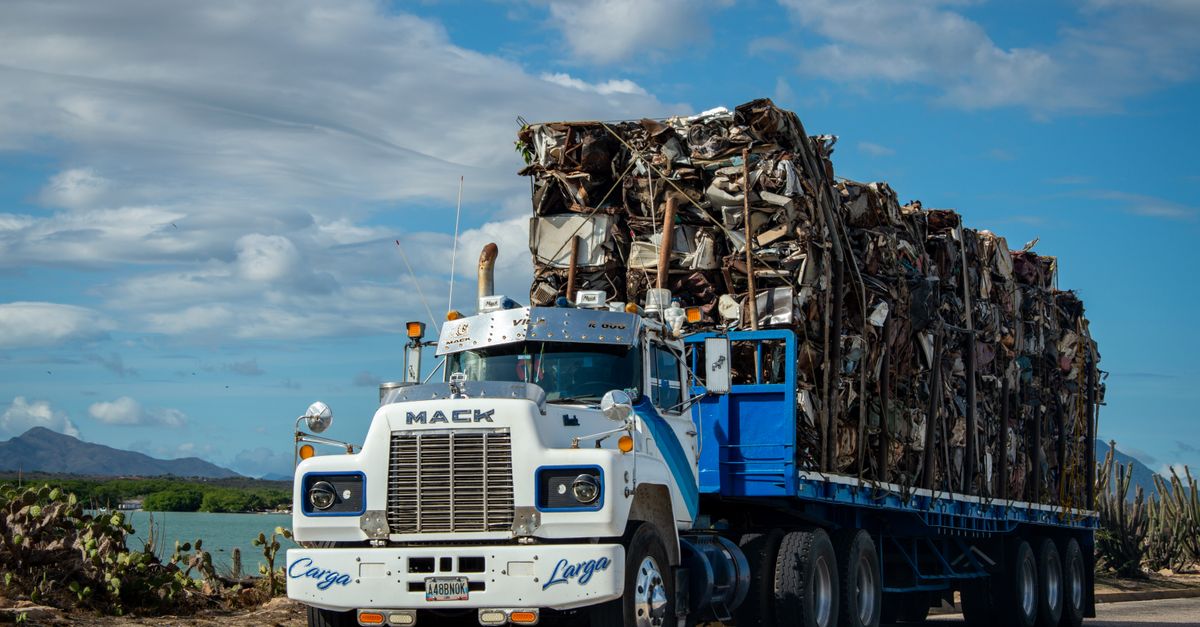 A Mack truck loaded with scrap metal travels on a rural road under a cloudy sky.