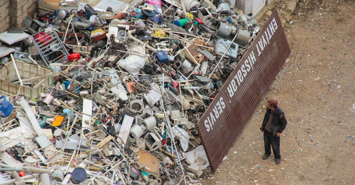 A person inspecting a large pile of scrap metal and electronic waste in an outdoor junkyard.