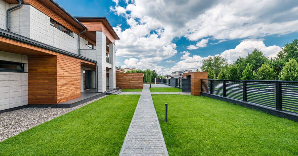 Scenery green yard of modern residential house with trees outside fence under blue cloudy sky