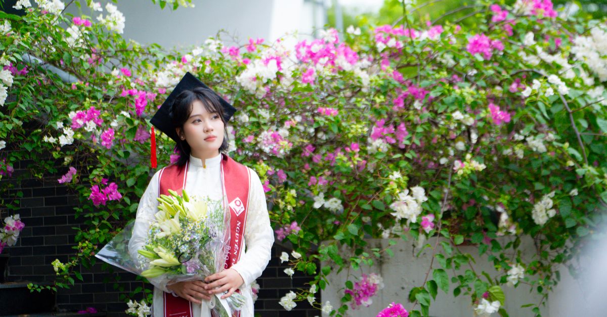 A young graduate in traditional attire holds a bouquet of flowers against a floral backdrop, celebrating her achievement.