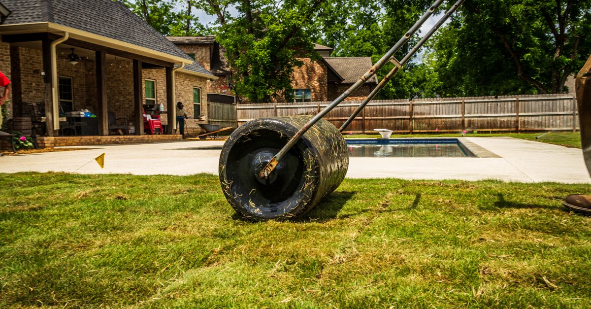 A worker rolling a lawn in a suburban backyard near a pool on a sunny day.