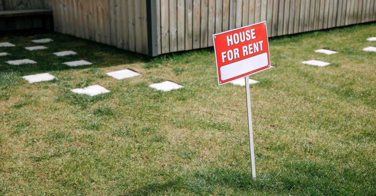 A red 'House for Rent' sign stands on a grassy lawn beside a wooden house exterior.