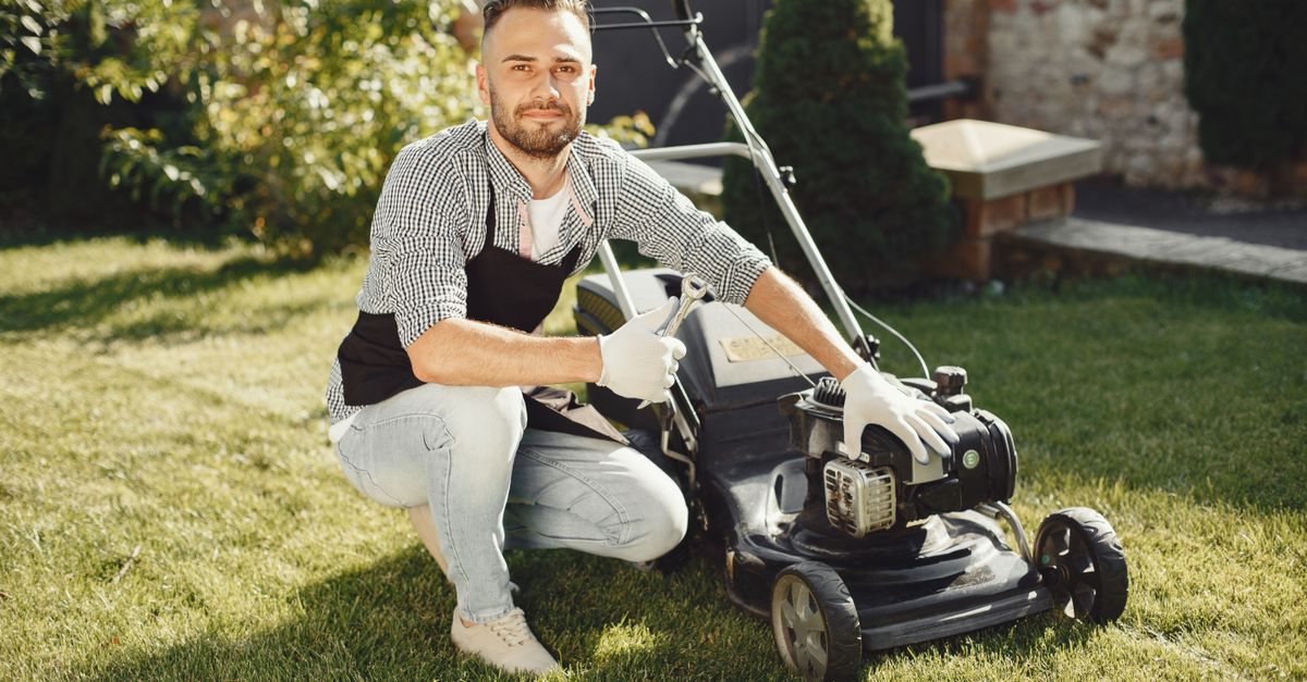 Adult man wearing apron and gloves maintains a lawn mower on a sunny day.