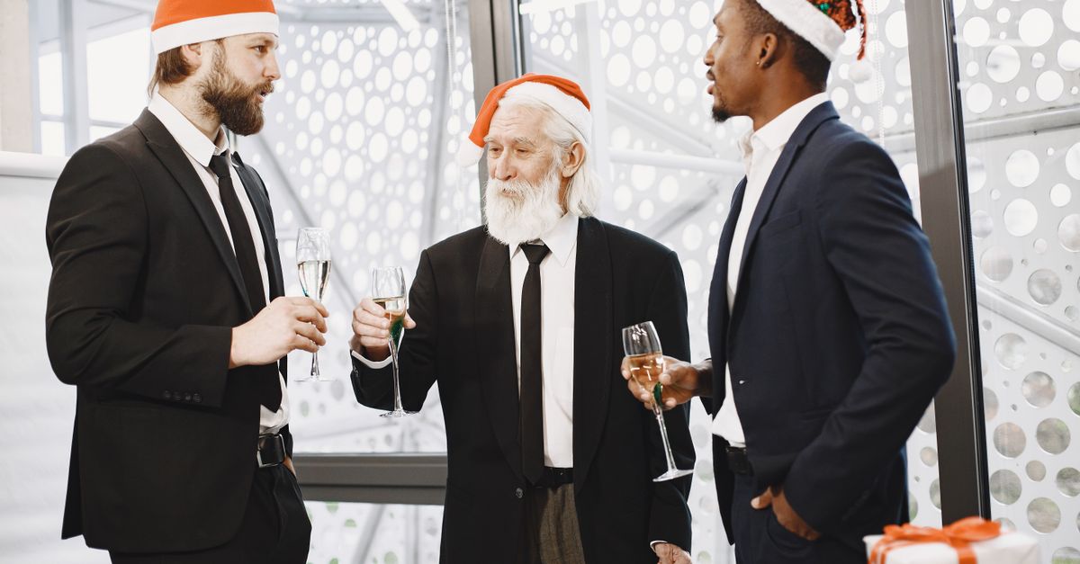 Three businessmen in suits celebrate Christmas with champagne indoors, wearing festive hats.