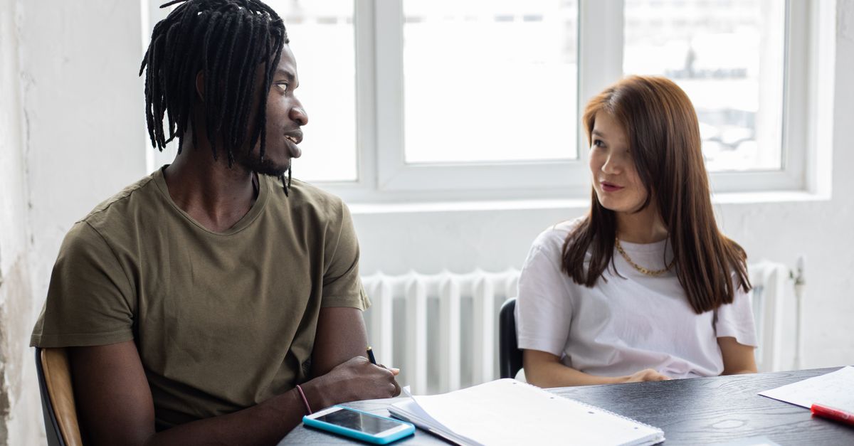 African American student and Asian female classmate chatting and looking at each other while sitting at table with notebooks during lesson