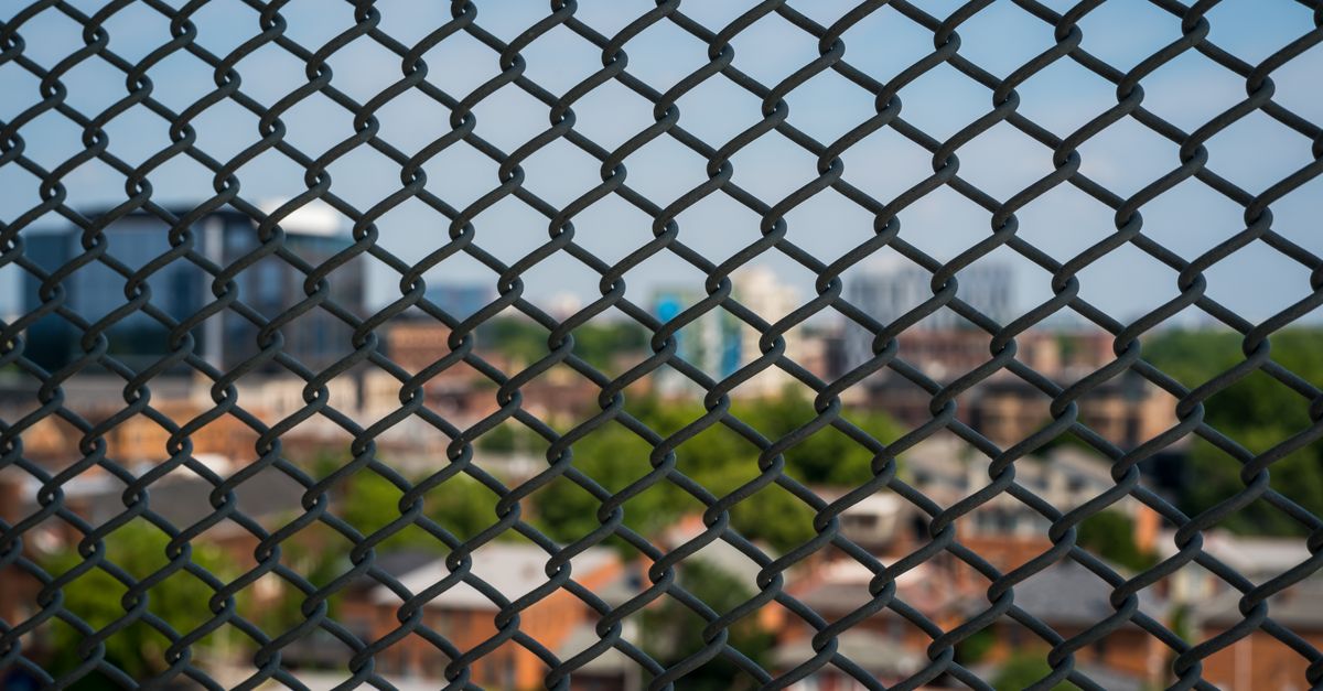 A cityscape blurred behind a metal mesh fence, showcasing urban architecture and greenery.