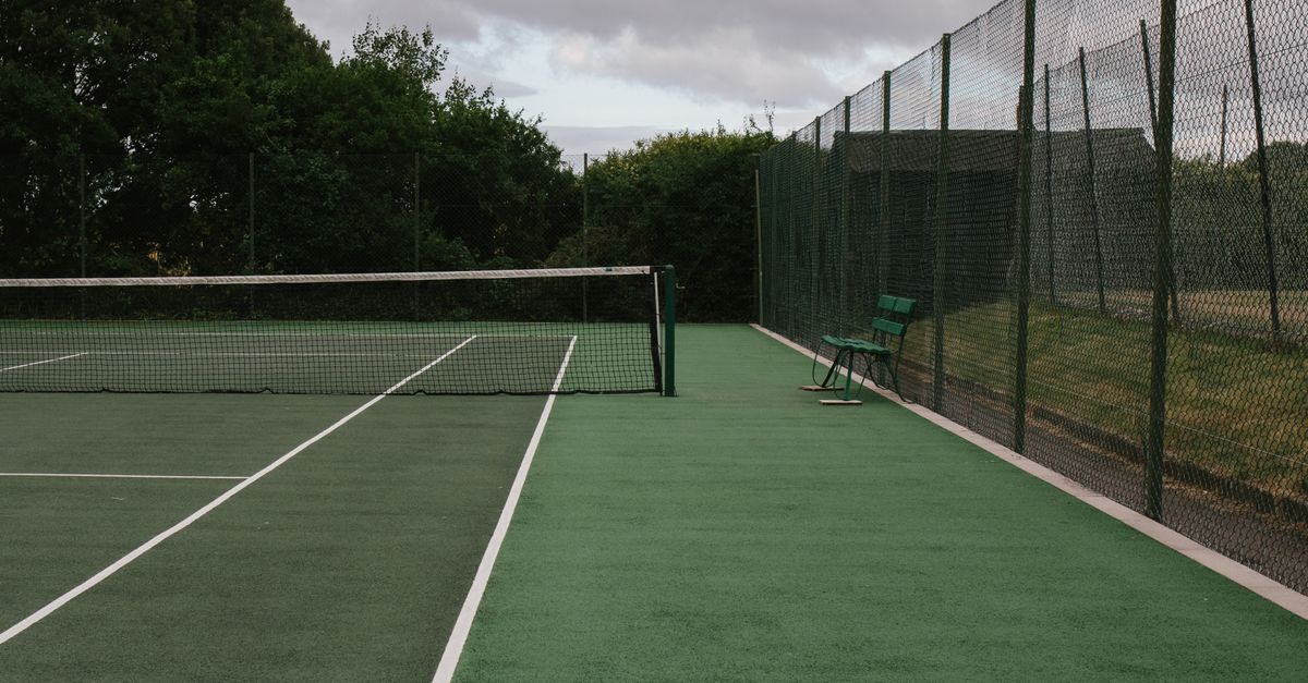 Green tennis court with net and fence under a cloudy sky, ideal for sports concepts.