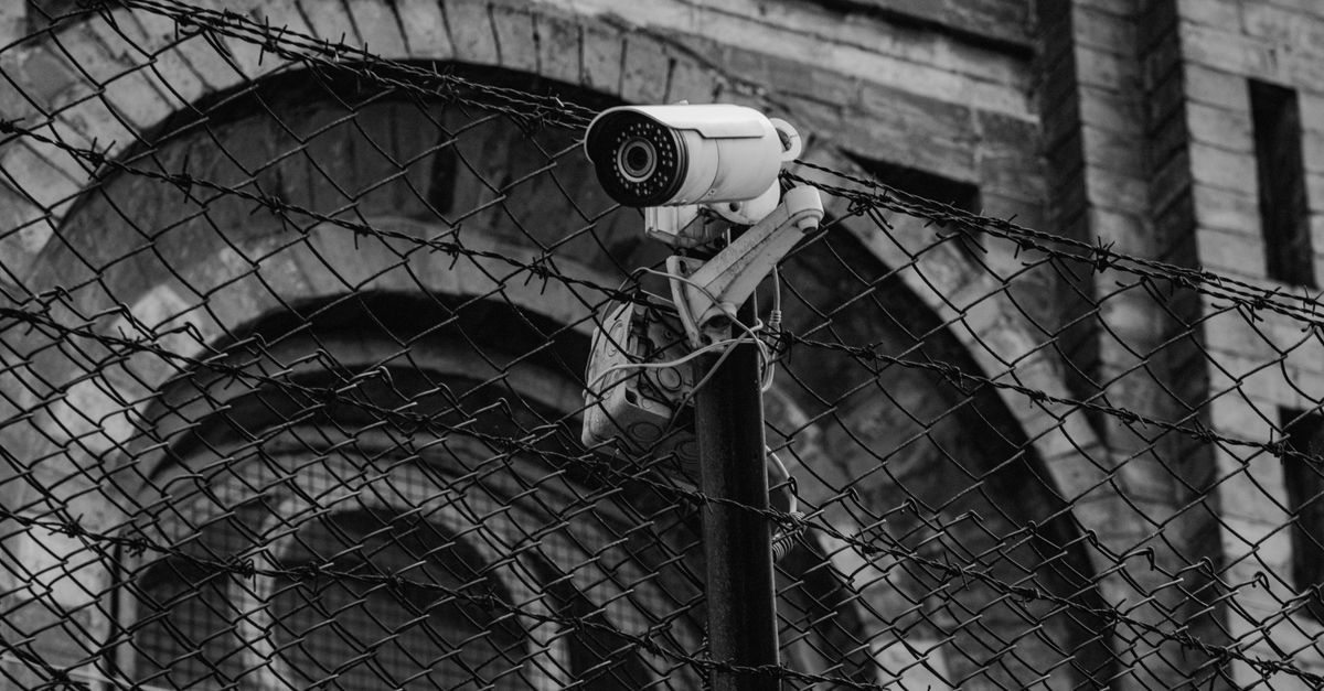 Black and white image of surveillance camera mounted on a fence outside a historic building.