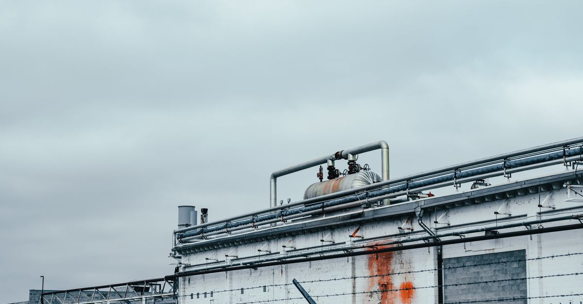 Shabby wall of factory with metal pipes and special equipment located near fence in industrial district of town against cloudy sky