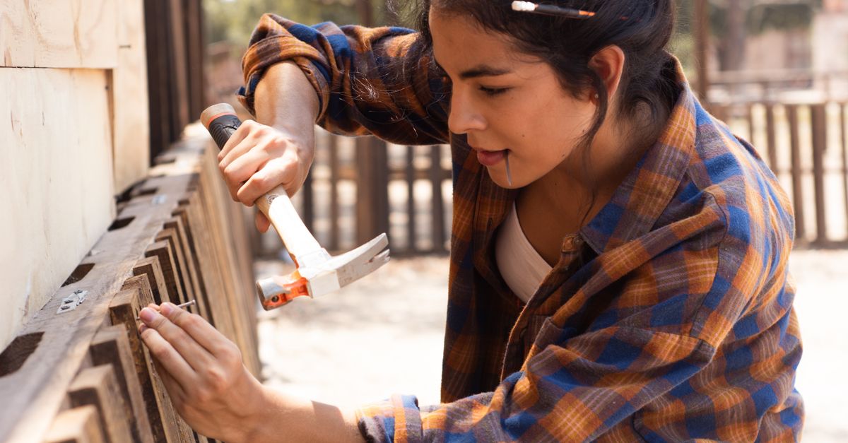 Young woman carpenter uses hammer to fix wooden fence outdoors.