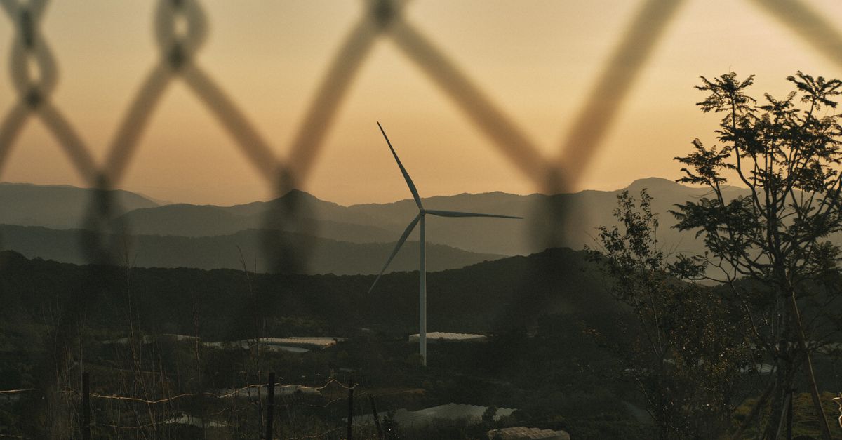 A wind turbine framed by a chainlink fence amidst mountains during sunset, symbolizing renewable energy.