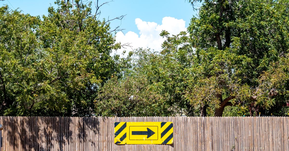 Yellow directional arrow sign on a wooden fence in a sunny outdoor setting.