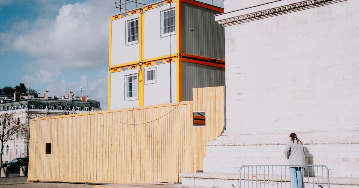 A woman walks by a construction site with stacked modular containers beside a classic building.
