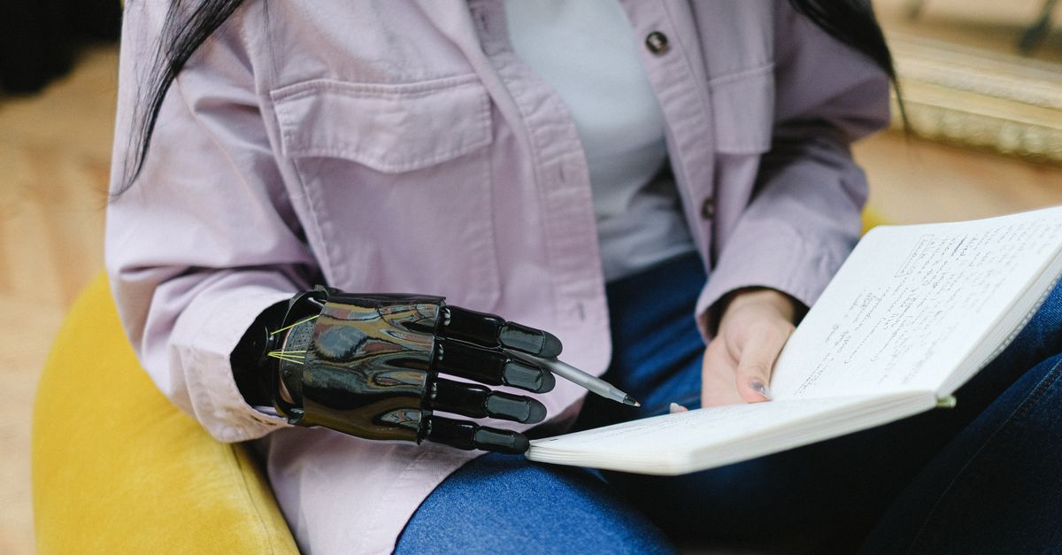 Close-up of a woman with a prosthetic hand writing in a notebook indoors, illustrating technology in daily life.