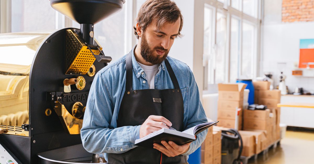 Man in denim shirt and apron writing notes beside a coffee roasting machine indoors.