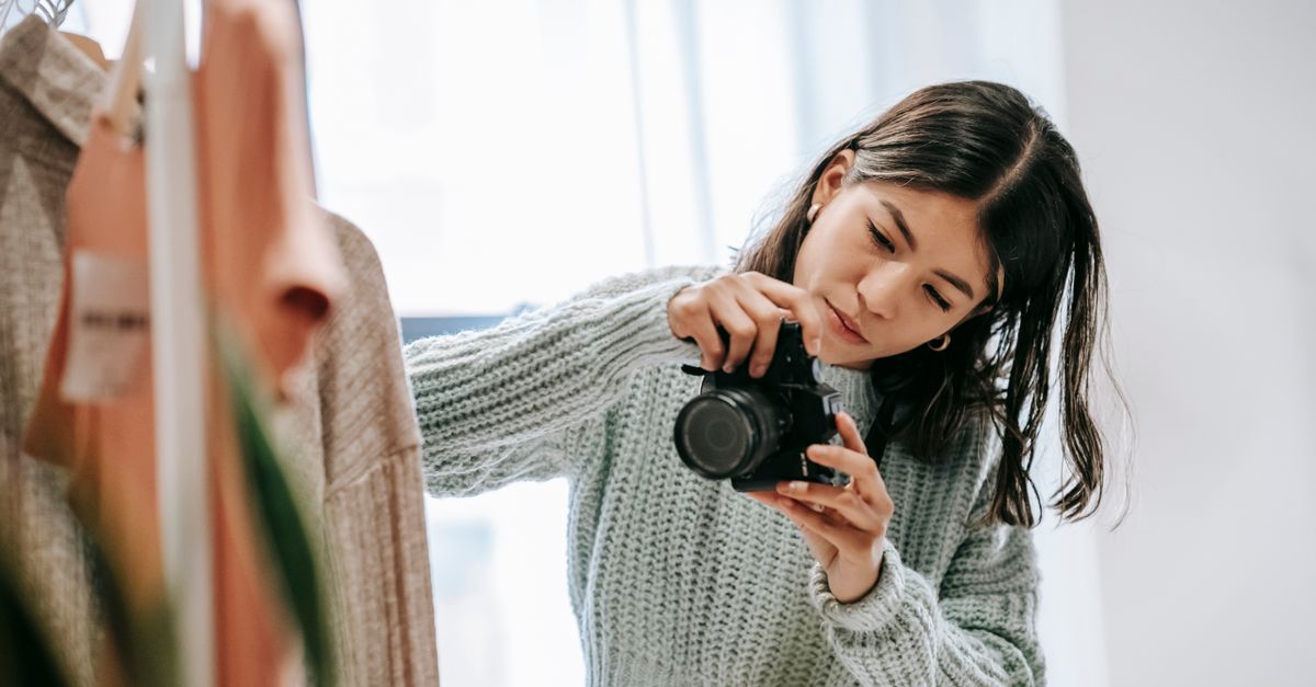 Young concentrated female photographer taking picture of clothes rack with hanging garment while looking at camera display in light room