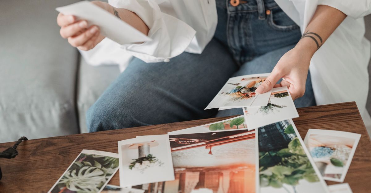 Crop unrecognizable tattooed female sitting on sofa at desk with assorted photos of flowers and town in house room