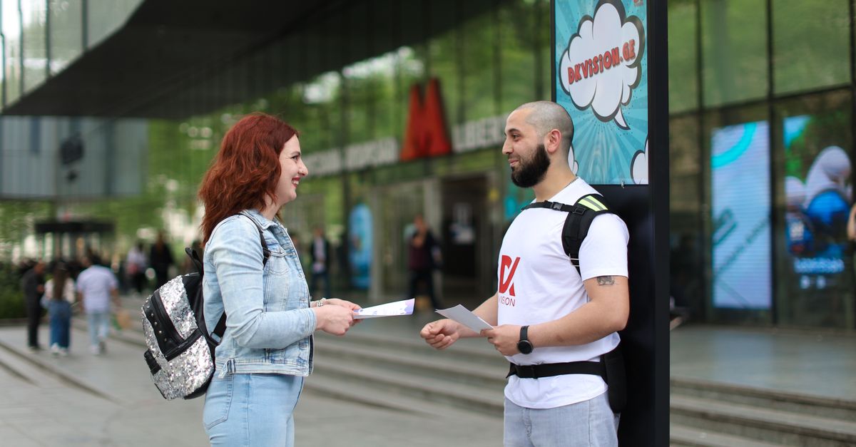 A man promoting with a backpack sign interacts with a woman on a busy city street.