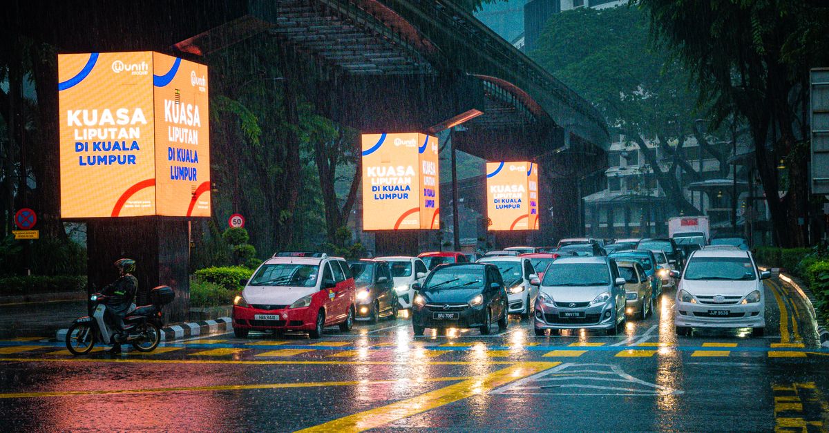 Traffic in Kuala Lumpur under heavy rain with illuminated advertisements and wet streets.
