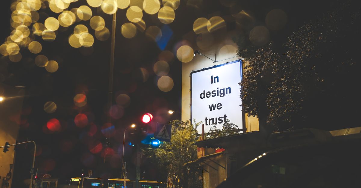 A city street at night featuring an illuminated billboard with the phrase 'In design we trust.'