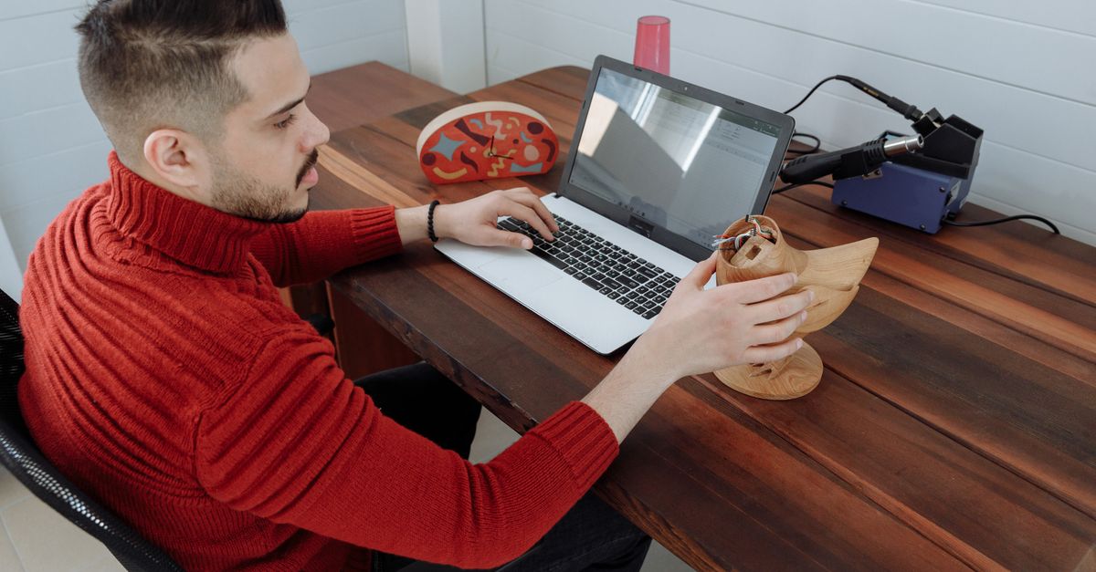 Engineer working on a laptop at a wooden desk, holding a 3D model in an office setting.
