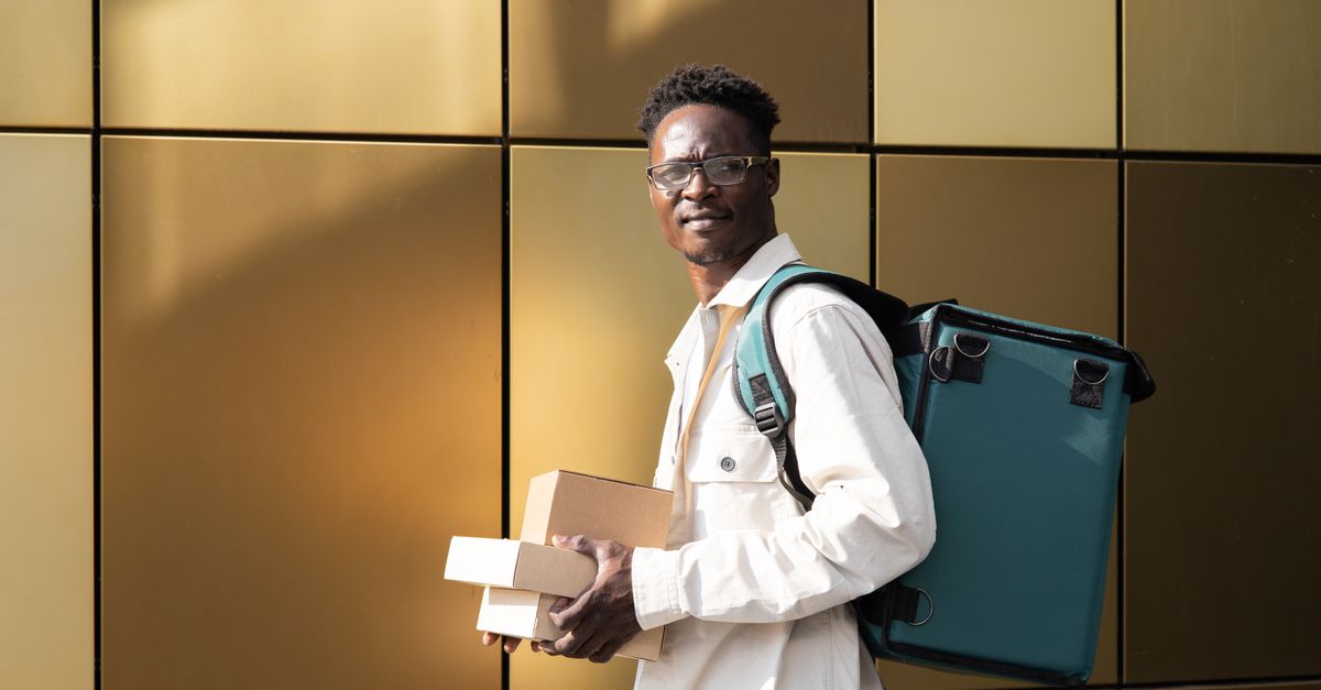 Young man with thermal bag delivering packages, wearing casual attire against gold wall.
