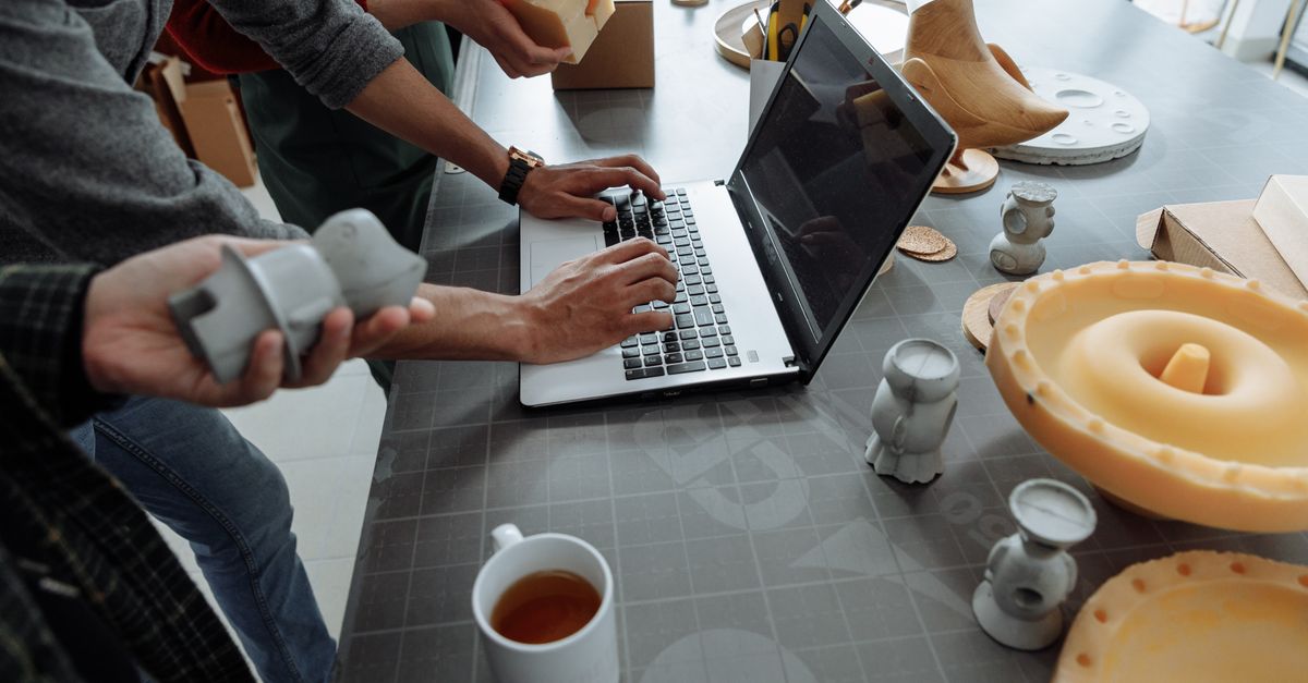 Hands on laptop in a creative workspace with 3D models and prototypes.