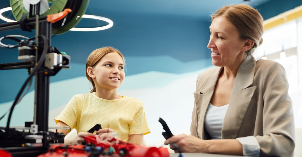 Low angle of smiling woman and little girl sitting at table and working on engineering project together while looking at each other in light workspace