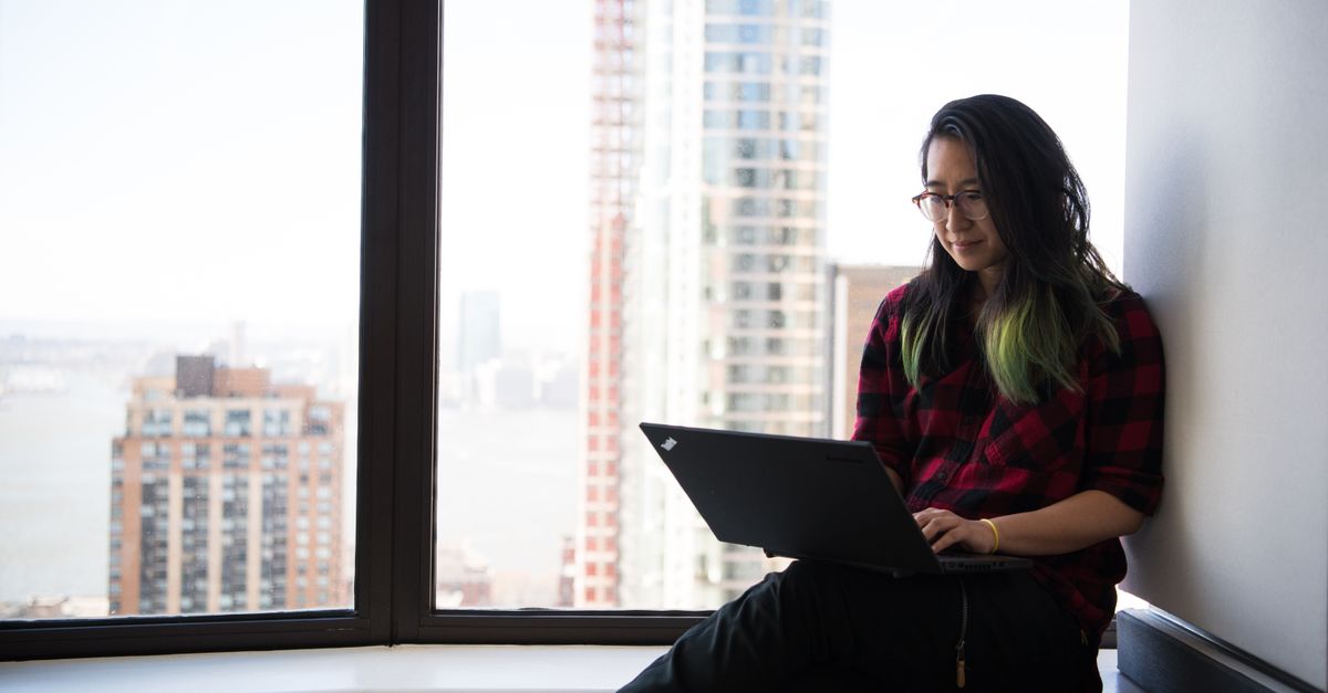 Young Asian woman working on laptop by city-view office window, embracing modern technology and workspace flexibility.
