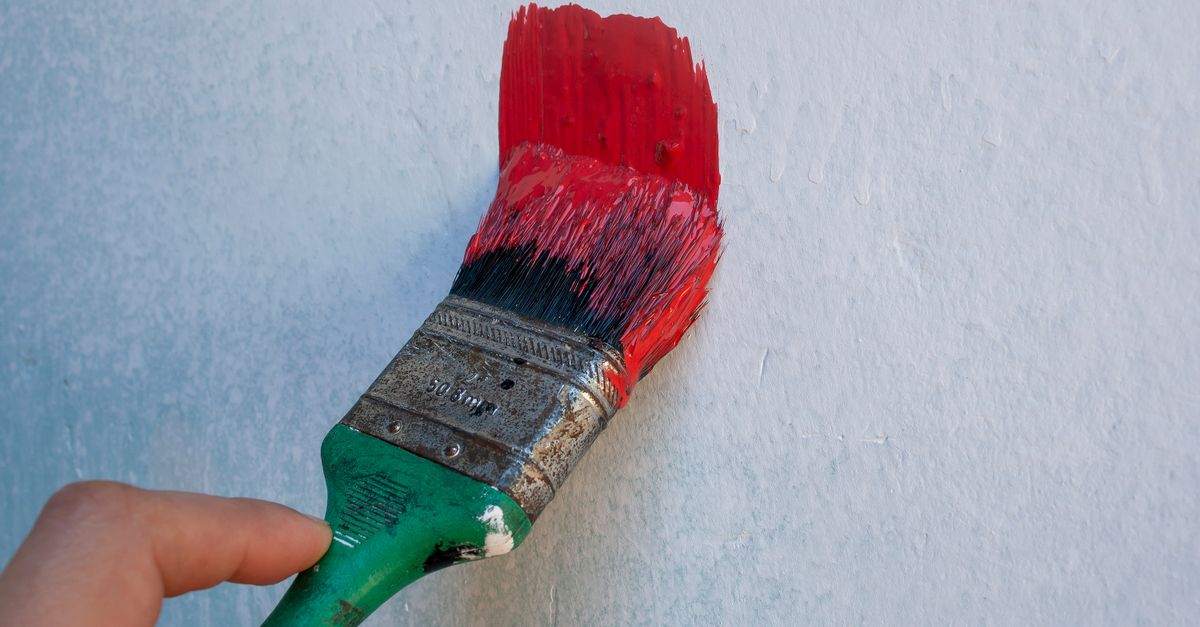 Detailed image of a hand holding a paintbrush with red paint on a textured wall.