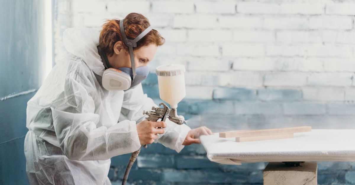 Woman in protective gear spray painting wood indoors against a brick wall.