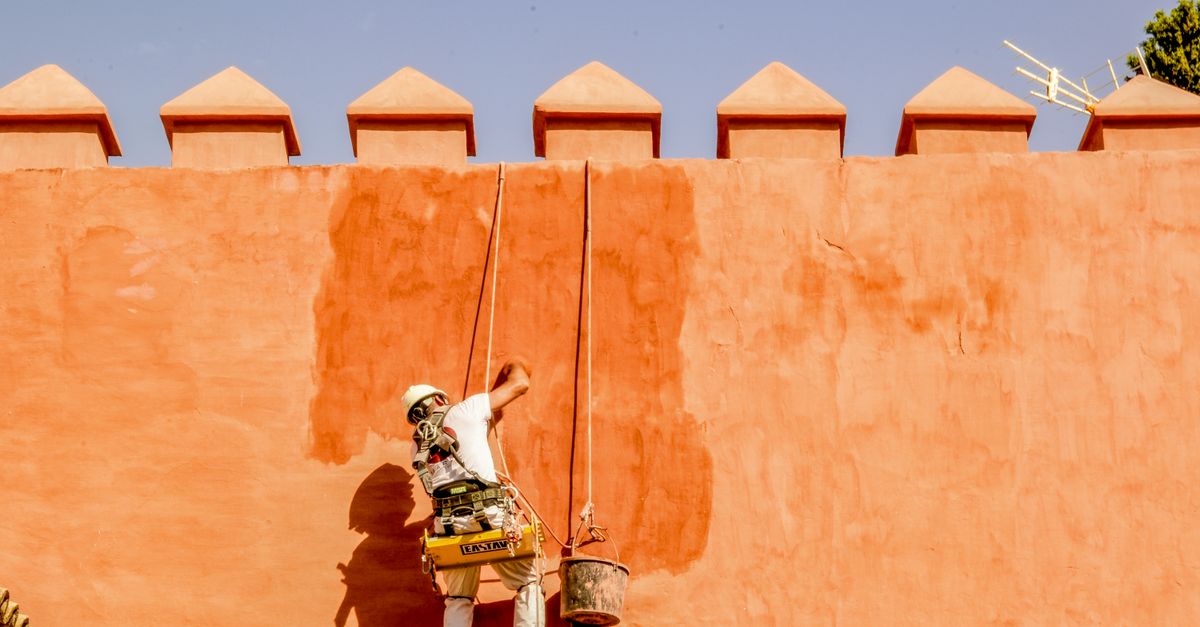 A worker repaints an orange brick wall in the sun, using ropes for support.