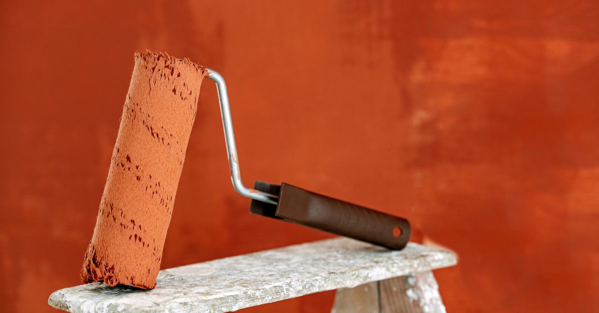 Detailed image of a paint roller resting on a ladder with a vibrant orange wall in the background.