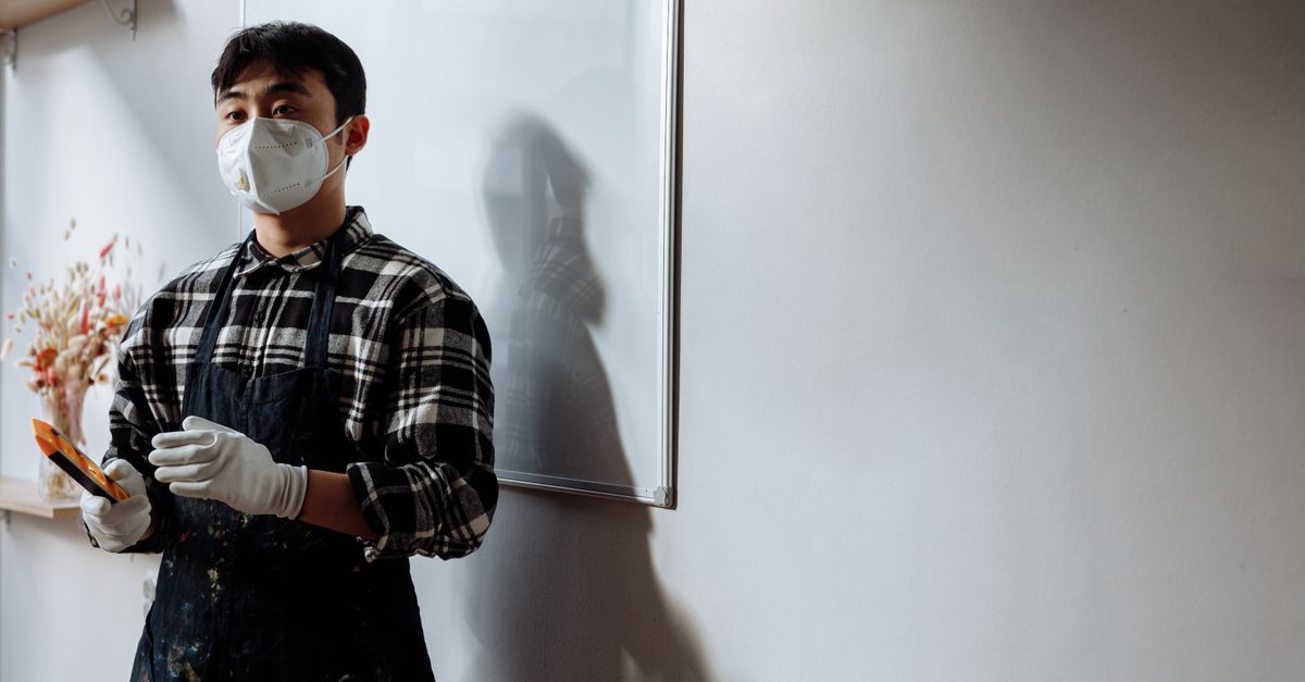 An artist wearing a face mask and apron holds brushes in a well-lit room with a whiteboard.
