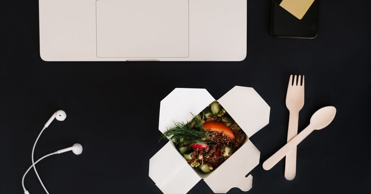 A flat lay of a workspace setup with a laptop, smartphone, earbuds, and a takeout meal.