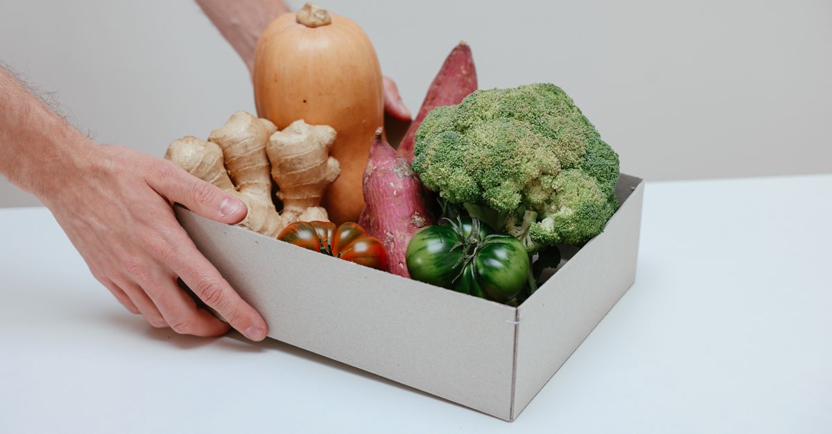 A cardboard box filled with fresh organic vegetables, being lifted by hands.